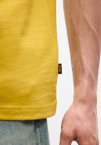 Άκρη κίτρινης μπλούζας με μικρή μαύρη ετικέτα "BOSS" που φορά άτομο με ανοιχτόχρωμο τζιν, εμφανίζοντας μ έρος του δεξιού βραχίονα και του χεριού.