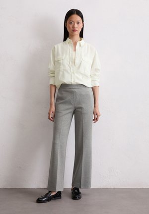 Lichtgele button-up shirt met twee zakken, gecombineerd met grijze, wijde broek. Het model staat met zwarte instappers op een neutrale achtergrond.