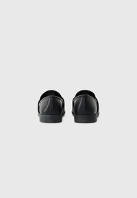 Pantofi slip-on din piele neagră, cu o textură netedă, formă rotundă, talpă joasă și detaliu cu logo embosat pe spate.