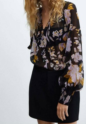 Femme portant un chemisier noir transparent avec un imprimé floral jaune et blanc et une jupe noire taille haute, debout devant un fond uni.