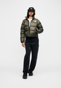 Γυναίκα που φοράει μπουφάν σε στυλ puffer σε ελιά, μαύρο καπέλο τύπου κάπ, μαύρο παντελόνι και μαύρα μποτάκια, μπροστά σε απλό λευκό φόντο.