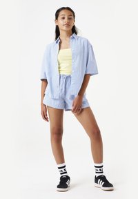 Blauw-wit gestreepte overhemden, gele tanktop, bijpassende gestreepte shorts, zwarte sneakers met witte accenten en gebloemde sokken.