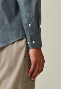 Chemise bleu foncé en tissu lisse, avec un poignet pointu, des boutons blancs et un ourlet légèrement incurvé. Associée à un pantalon beige clair.