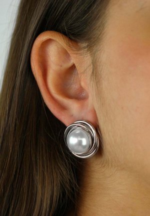 Oreja con un gran pendiente de plata que presenta una perla central rodeada por bandas metálicas circulares entrelazadas, llevado en el lóbulo izquierdo.