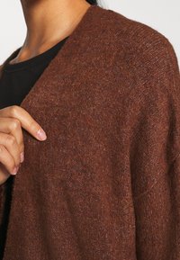 Braune Strickjacke mit weicher Textur, offenes Design an der Vorderseite und weitem Ärmel, die eine lockere Passform und subtile Farbvariationen zur Schau stellt.