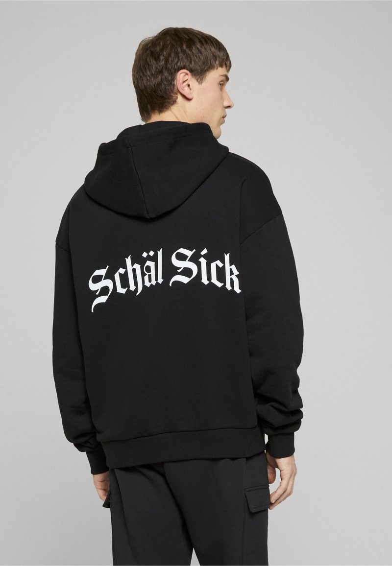 Jeune homme portant un sweat à capuche noir avec l'inscription blanche "Schäl Sick" dans le dos, se tenant de profil devant un fond gris uni.