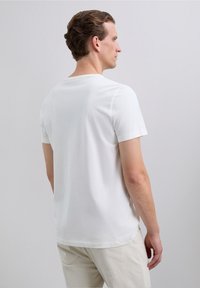 Uomo con i capelli castani corti che indossa una maglietta bianca a maniche corte e pantaloni crema, in piedi di lato contro uno sfondo grigio chiaro.