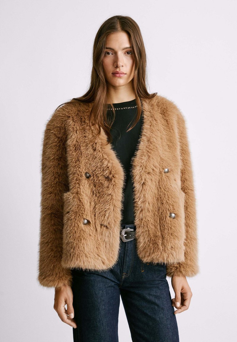 Braune Kunstpelzjacke mit doppelter Knopfleiste, zwei vorderen Taschen und silberfarbenen Knöpfen, kombiniert mit einem schwarzen Oberteil und Jeans.