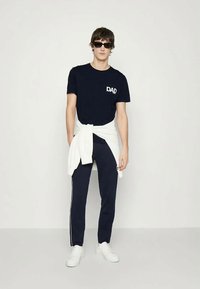 Jauns vīrietis ar tumšām saules brillēm, valkājot melnu krekliņu ar uzrakstu "DAD", tumši zilas bikses ar baltām svītrām, baltas sporta kedas un baltu džemperi sasietu ap vidukli.