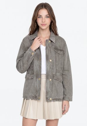 Junge Frau mit langen braunen Haaren, die Creolen trägt, eine graue, geknöpfte Jacke, ein weißes Top und einen beigen plissierten Rock, stehend vor weißem Hintergrund.