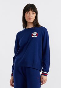 Blauwe wollen trui met ronde hals, geborduurd logo met rode en witte accenten en gestreepte manchetten in rood, wit en blauw.
