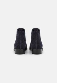 Un paio di stivali alla caviglia in suede blu scuro con tacco basso, visti da dietro su uno sfondo grigio chiaro uniforme.