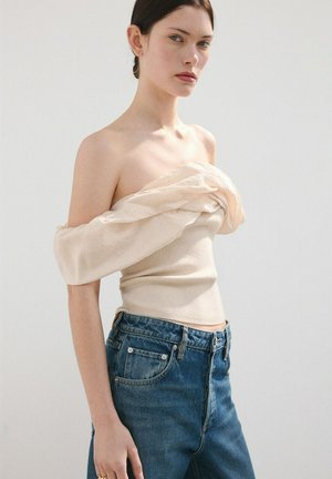 Joven con cabello oscuro recogido lleva una blusa beige de hombros descubiertos y jeans de mezclilla azul de cintura alta contra un fondo liso.