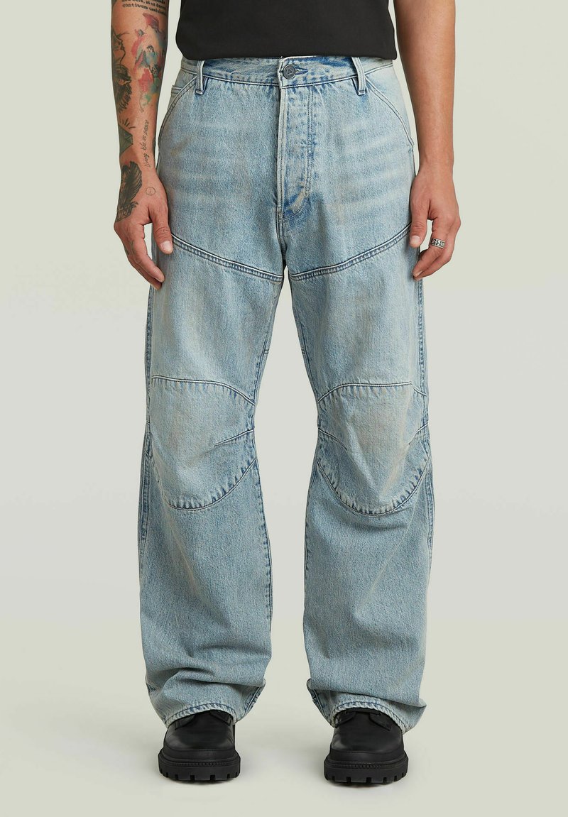 Jeans di denim azzurro chiaro con dettagli a toppe sulle ginocchia, indossati da una persona con tatuaggi, scarpe nere e top nero su sfondo semplice.