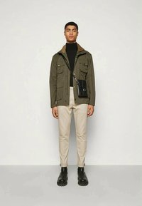 Young man wearing an olive jacket, black turtleneck, beige pants, black boots, and a black crossbody bag, standing against a plain background.