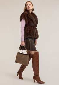 Kobieta w brązowej futrzanej kamizelce, różowym swetrze, brązowej skórzanej spódnicy, brązowych kozakach za kolano, z taupe torebką, pozująca na jednolitym tle.