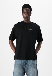 Camiseta de algodón negra con mangas cortas y cuello redondo. Texto "atelier dept." en dorado en el pecho. Combinada con jeans de mezclilla de color azul claro.