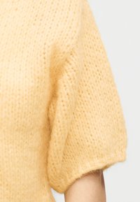 Κοντινή λήψη ενός μαλακού, χνουδωτού, ανοιχτού κίτρινου πλεκτού μανικιού που καλύπτει το πάνω μέρος του βραχίονα ενός ατόμου σε λευκό φόντο.