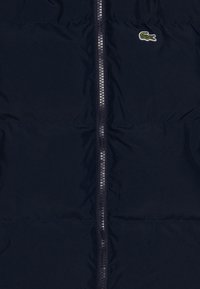 Marineblå puffjakke med en sentral glidelås. Har et lite brodert krokodillelogo. Laget av glatt, lett stoff med quiltet tekstur.