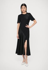 Fekete ruha testhez simuló felsőrésszel és bővülő szatén szoknyával. Rövid ujjakkal, aszimmetrikus dizájnnal és combig érő hasítékkal díszített.