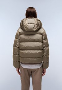Persona di spalle che indossa una giacca a vento imbottita color verde oliva e pantaloni marroni su uno sfondo grigio chiaro.