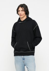 Sort hoodie med grå syninger, en frontlomme og hætte med snøre, båret over lyseblå denim jeans. Blødt stof, afslappet pasform.