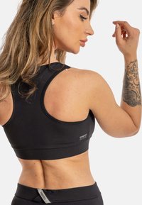 Svart sports-bh laget av elastisk materiale, med scoop-back design og en liten logo nederst. Glatt tekstur og tettsittende form.