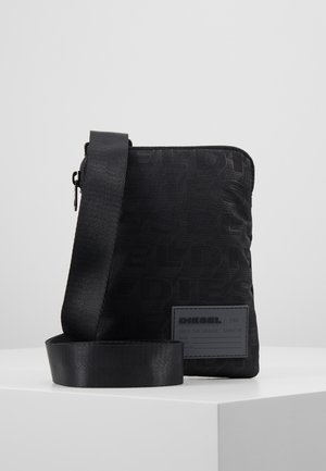 Kleine schwarze Diesel Crossbody-Tasche mit verstellbarem Riemen und Logo-Patch vorne, stehend auf einer weißen Oberfläche vor einem einfarbigen Hintergrund.