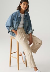 Giubbotto in denim dalla vestibilità rilassata, di colore azzurro chiaro, con maniche ripiegate; abbinato a pantaloni beigi a pieghe e sneakers bianche. Seduto su uno sgabello di legno.
