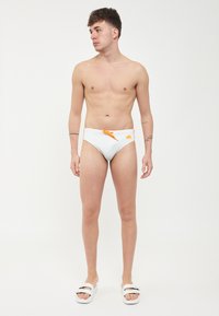 Des slips de bain blancs avec une touche orange, une texture lisse et un design taille basse. Portés avec des sandales blanches à glisser.
