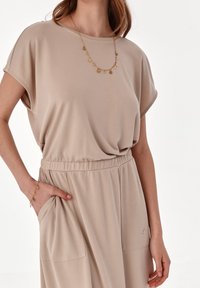 Béžové šaty s krátkým rukávem, volného střihu, s elastickým pasem, kapsami a dekoračním zlatým náhrdelníkem s kulatými přívěsky.