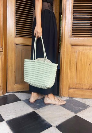 Persoană îmbrăcată într-o rochie neagră, ținând o geantă de umăr împletită de culoare bej deschis, purtând pantofi loafer bej, stând pe un podea în carouri alb-negru lângă uși din lemn.