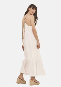 Vestido maxi blanco con detalle de lazo en la espalda, dobladillo acampanado y tirantes finos, combinado con sandalias de cuña beige con tiras entrecruzadas.