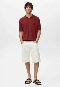 Red short-sleeve polo shirt, textured fabric, paired with cream shorts. Black sandals with a buckle detail. Simple, casual outfit.