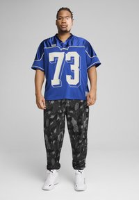 Jersey de malla azul con el número blanco "73", combinado con pantalones de chándal con patrón de camuflaje negro. El modelo lleva zapatillas deportivas blancas. Fondo sencillo.