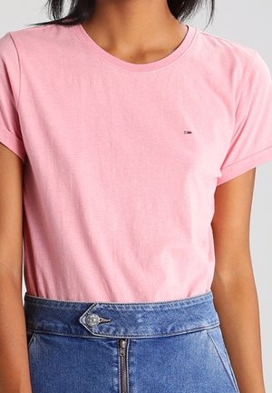 Osoba nosząca jasnoróżowy t-shirt z krótkim rękawem wpuszczony w niebieskie, wysokie dżinsy z przodu na zamek błyskawiczny i guzik.