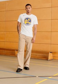 Hvid t-shirt med "Golden State Warriors" logo, beige stribede bukser og sorte sko; stående på et gymnastikgulv med trægulve.