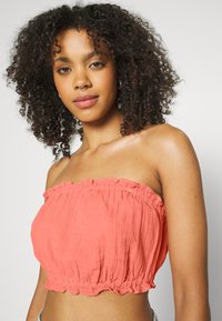 Top cropped sem alças em coral, confeccionado em tecido leve com textura franzida e bainha com babados. Apresenta um acabamento suave e um caimento descontraído.