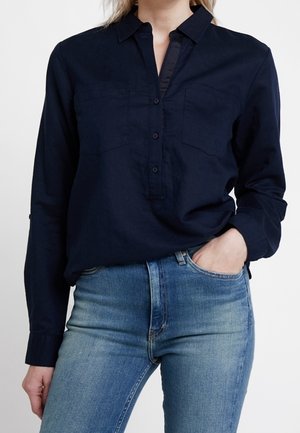 Kvinde iført en mørkeblå langærmet skjorte med knapper, stoppet ned i lyseblå højtaljede jeans, med den ene hånd i lommen.