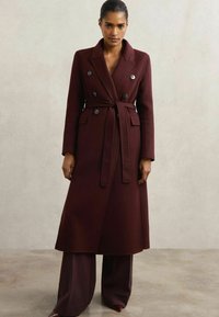 Mørkerød uld lang frakke med dobbeltradede knapper, bæltebånd og forlommer. Glat overflade, skræddersyet form og sømfaldsdesign.