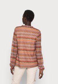 Blusa de mangas longas com padrão em zig-zag nas tonalidades de laranja, rosa e castanho; textura translúcida com punhos franzidos; decote redondo.