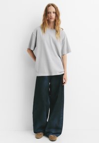 Tricou oversized gri deschis cu mâneci scurte, asortat cu blugi tăiați larg din denim albastru închis și saboți maro, cu un model. O ținută simplă și relaxată.