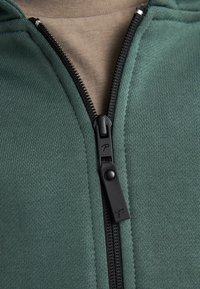 Groene zip-up jacket van een textuurstof, voorzien van een zwarte rits en een beige binnenlaag. Het ontwerp bevat geribbelde details.