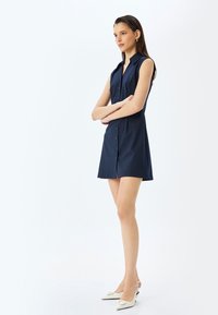 Navy blue ilmanhiha dress where the front is adorned with buttons, featuring an A-linjainen siluetti and sivureiät. Käytetään valkoisten slingback-korkokenkien kanssa. Sujuva kangastuntuma.