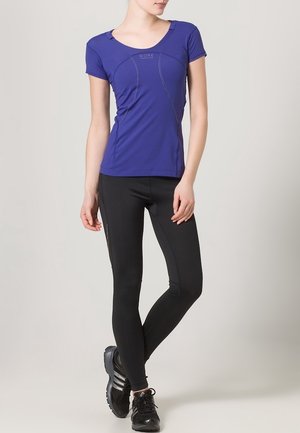 Lila kurzärmeliges Sportshirt mit geschwungenen Nähten, kombiniert mit schwarzen enganliegenden Leggings. Das Modell trägt schwarze Sneakers. Einfaches Design, leichtes Material.