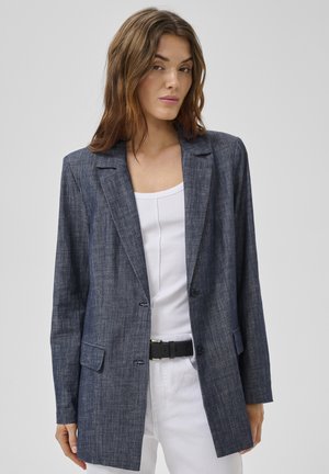 Kvinde med bølget brunt hår iført en mørkegrå blazer over en hvid tanktop og hvide bukser med sort bælte, stående mod en ensfarvet baggrund.