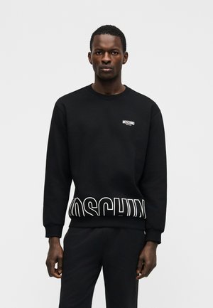 Homme debout, portant un sweat-shirt noir Moschino avec le logo blanc sur la poitrine et une grande impression du logo en bas.