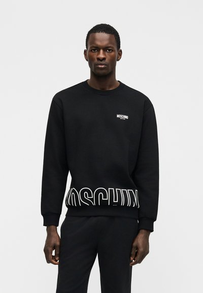 Άνδρας που στέκεται, φορώντας μαύρο φούτερ Moschino με λευκό κείμενο λογότυπου στο στήθος και μεγάλο λογότυπο στο κάτω μέρος.