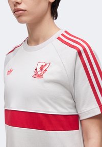 Άτομο που φοράει ανοιχτό γκρι μπλουζάκι Adidas με κόκκινες ρίγες στον ώμο και το έμβλημα της Liverpool Football Club στο στήθος.