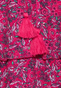 Tissu rose vif avec des motifs floraux en vert, blanc et noir, comportant deux pompons roses suspendus avec des sommets ronds.